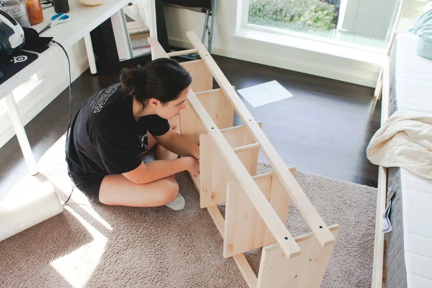 Someone sitting on a rug assembling a wooden shelving unit in a sunny room.